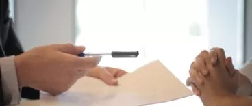 person handing pen and paper to someone sitting across from them with their hands folded